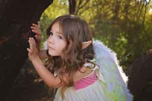 Little girl with long brown hair, pointed elf ears and pastel fairy wings peers around a tree trunk in a sunlit forest.