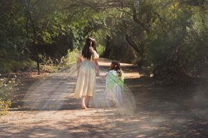 A woman in a cream dress walks barefoot with a little girl in green fairy wings along a sunlit wooded forest path.