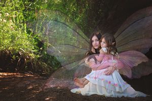 A woman and a young girl wearing delicate fairy wings sit close together on the forest floor in soft dappled sunlight.