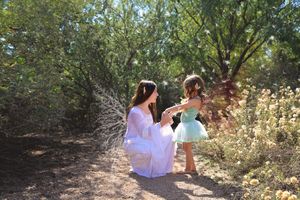 A woman in a flowing white gown kneels and holds hands with a little girl in a teal fairy dress beside blooming wildflowers.