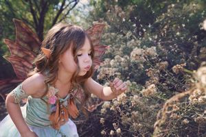 A young girl with pointed fairy ears and wings gently blows on delicate wildflowers in a sparkling, magical forest scene.