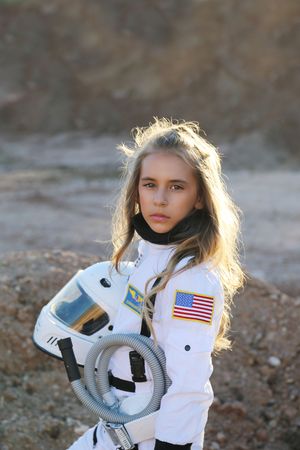 A young girl in a white astronaut suit with an American flag patch holds her helmet against a rocky, Mars-like landscape.