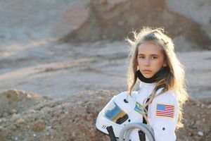 A serious young girl with windswept blonde hair wears a white NASA-style spacesuit and cradles her helmet in a rugged desert.