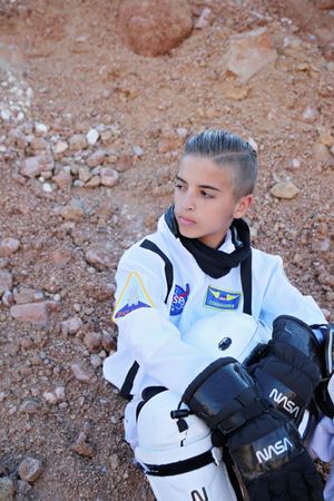 A young astronaut in a white spacesuit with NASA gloves sits on red rocky terrain, gazing off to the side in a Mars-like setting.