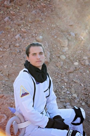 A young man in a white astronaut suit and black neck gaiter sits on rocky red ground in a Mars-like desert landscape.