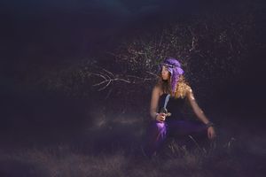 A woman in a purple outfit and headscarf crouches in a dark, moody field holding a dagger and looking off to the side.