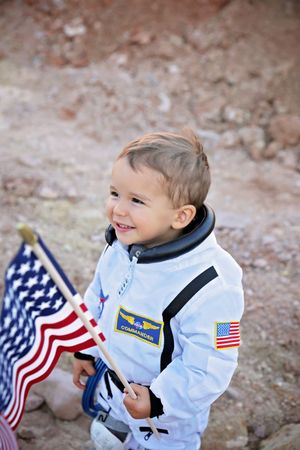 Smiling toddler in a white astronaut costume holds a small American flag on rocky, reddish terrain.