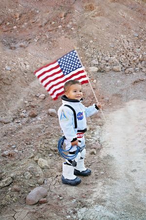 Toddler in a NASA-style astronaut suit waves an American flag while standing on rugged, rocky ground.
