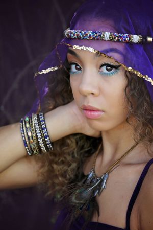 Close-up portrait of a woman with curly hair, blue eyeshadow, a purple veil, beaded headband, stacked bangles and feather necklace.