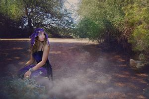 Woman in a purple veil and gown sits in a hazy wooded clearing surrounded by green trees and drifting mist.