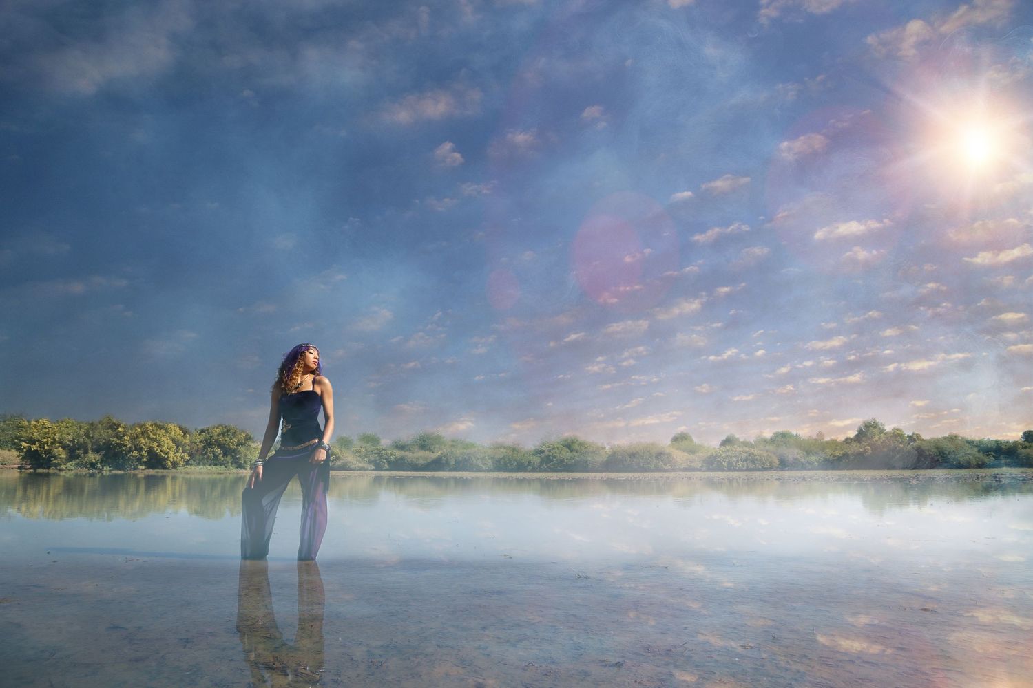 Woman stands in shallow water under a dreamy blue sky with glowing sun and soft clouds reflecting on the surface.