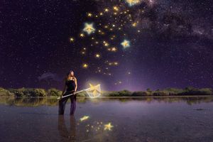 Woman standing in shallow water at night holding a wand that sends glowing golden stars into a purple Milky Way sky.