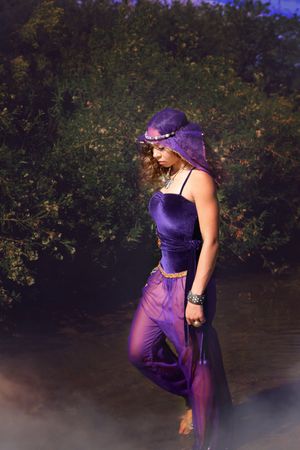Woman in a purple velvet gown and veil walks barefoot beside dark water with lush green foliage and mist behind her.