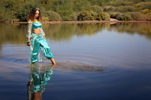 A woman in a turquoise princess-style costume wades barefoot through calm lake water, her reflection rippling below.