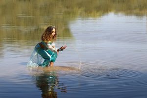 A curly-haired woman in a teal costume kneels in shallow lake water, splashing droplets as ripples spread around her.