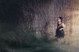 A young girl in a dark dress holds a doll beside a weathered lattice fence in a moody, misty field at dusk.