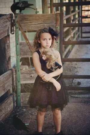 A serious young girl in a black tulle dress hugs a doll beside a rustic wooden gate while crows perch nearby.