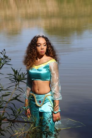 A woman in a satin turquoise princess costume and tiara stands at the lake shore beside green leaves in soft sunlight.