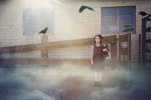 A girl in a dark dress holding a doll stands in a hazy farmyard as crows fly and perch around a rustic barn.