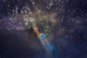 Dreamy fantasy portrait of a woman in turquoise costume gazing upward beneath a tree in misty, glowing purple light.