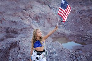 A girl in a white and blue astronaut-inspired costume holds up a waving American flag among rugged red rocks and a small pool.