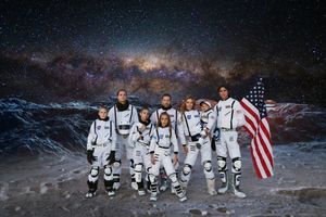 Family of seven in white astronaut suits poses on a rocky lunar surface holding an American flag beneath a starry galaxy sky.