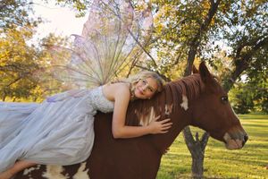 A smiling girl in a pale gown with sparkling fairy wings rests across the back of a paint horse beneath orchard trees.