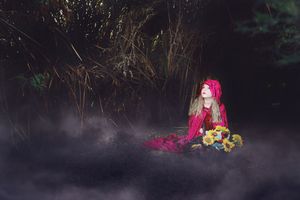 Girl in a red hooded cloak sits on the ground holding sunflowers in a dark misty forest.
