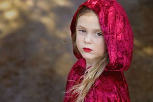 Young girl with long blonde hair and red lipstick wearing a crimson velvet hooded cloak against a softly blurred outdoor background.