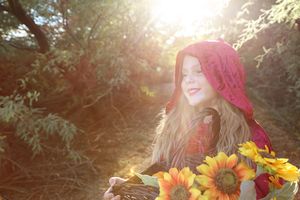 Smiling girl in a red hood holds a basket of sunflowers as golden sunlight streams through the trees.