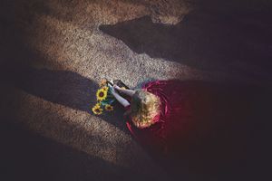 Overhead view of a girl in a red cloak sitting on sandy ground with sunflowers and long dramatic shadows.