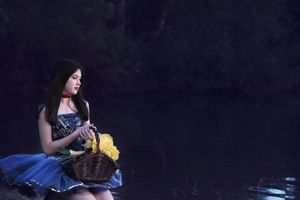 Girl in a blue dress with a red choker sits by dark water holding a basket of yellow flowers.