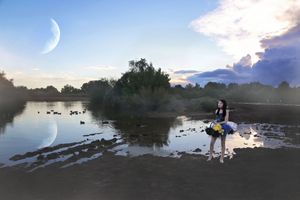 Girl in a tutu stands beside a calm pond at twilight with a crescent moon reflected in the water.