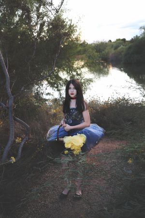 Young girl in a blue tulle skirt sits by a tranquil lake at sunset holding a basket of yellow roses.