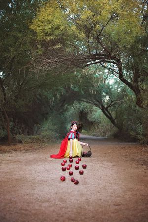 Girl dressed as Snow White sits on a forest path as red apples spill from her basket onto the ground.