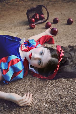 Girl in a blue and red princess costume lies on the ground with red apples spilling from a woven basket nearby.