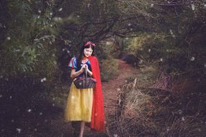Woman in a Snow White-style costume with red cape and yellow skirt holding a basket on a forest path as snow falls.