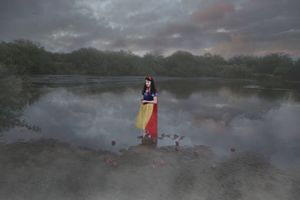 Person in a Snow White-style costume with yellow and red skirt standing on a muddy shore beside a calm lake under a moody cloudy sky.