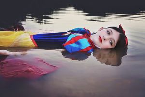 Girl in a Snow White costume with red lips floats on her back in calm water, reflecting the sky.