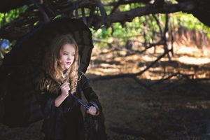 A young girl with long wavy hair stands under a black lace parasol wearing a dark gothic dress among shaded trees.