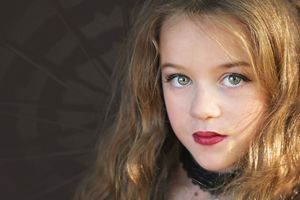 Close-up portrait of a young girl with wavy auburn hair, green eyes and dark red lipstick against a dark background.