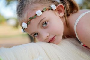 A young girl with a white flower crown rests her head on a white horse's neck in a soft, gentle outdoor portrait.