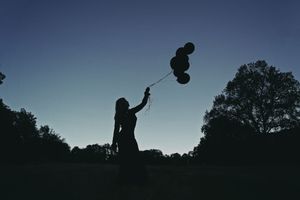Silhouette of a woman holding a bunch of black balloons against a deep blue dusk sky with dark trees behind her.