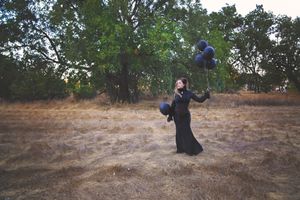 A woman in a long black dress stands in a dry grass field holding black balloons beneath large green trees.