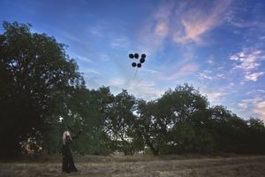 A woman in a dark dress releases black balloons into a blue sky with pink clouds above a row of oak trees.