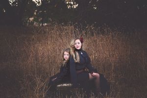 Two girls in dark gothic dresses sit together in a golden autumn field at dusk in a moody portrait.