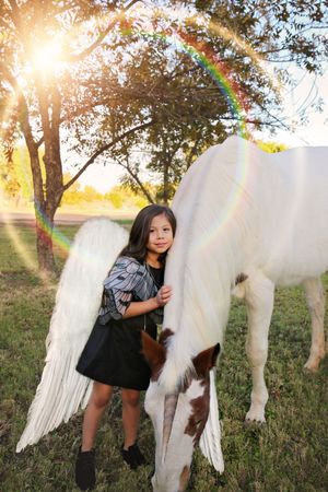 A girl with white angel wings stands hugging a white and brown horse under a sunlit tree with a rainbow lens flare.