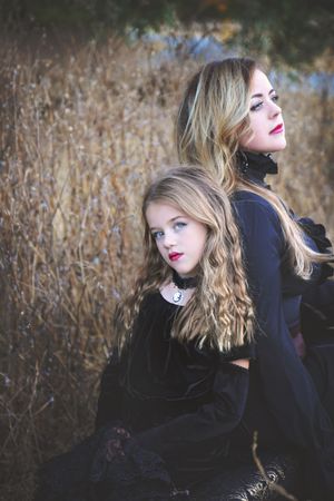 Two girls in black velvet gothic outfits with red lipstick pose together against dry golden grass in an autumn portrait.