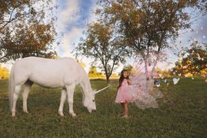 Little girl in a pink fairy costume stands before a white unicorn grazing in a grassy meadow at golden hour.