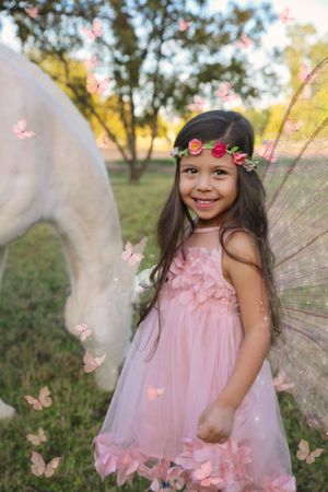 Smiling girl in a pink fairy dress with floral crown and wings stands near a white unicorn as butterflies flutter around.